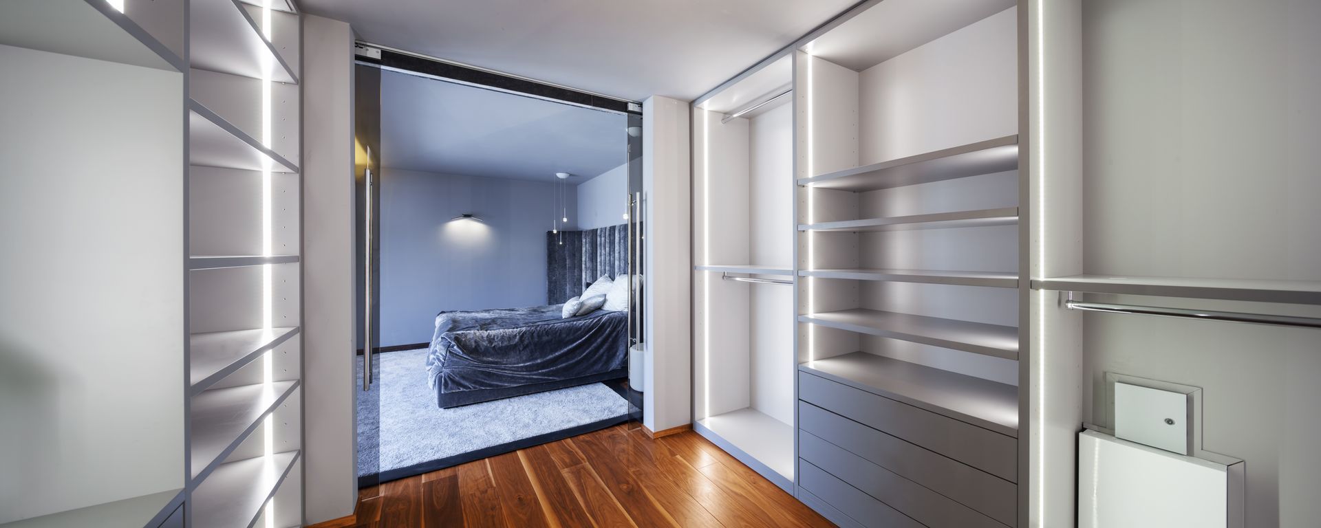Dressing avec étagères, tiroirs et tringles à vêtements. La porte ouverte donne sur une chambre avec un lit.