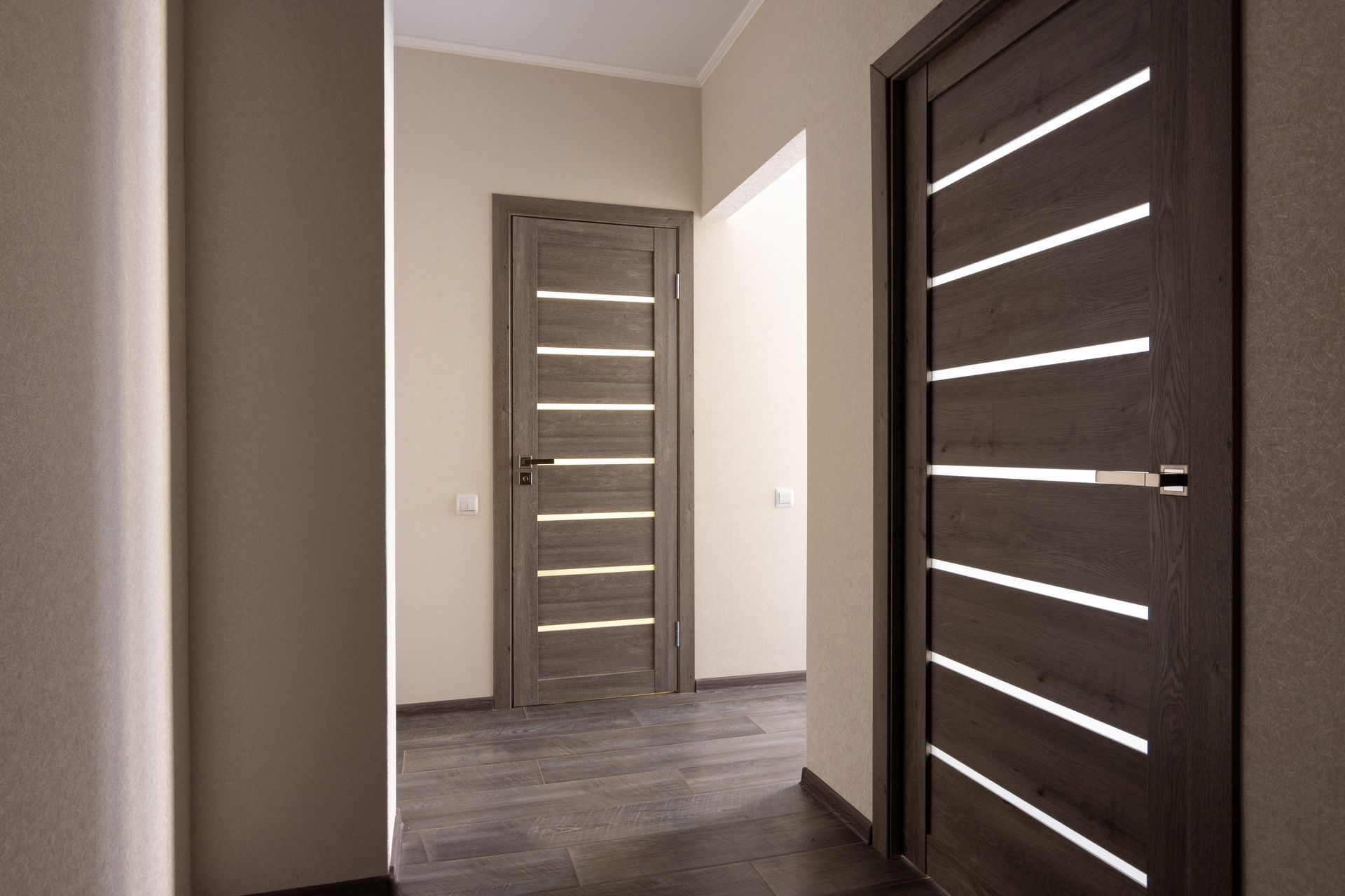 Couloir avec trois portes : deux portes grises fermées avec des fentes horizontales, une ouverte sur une pièce blanche.