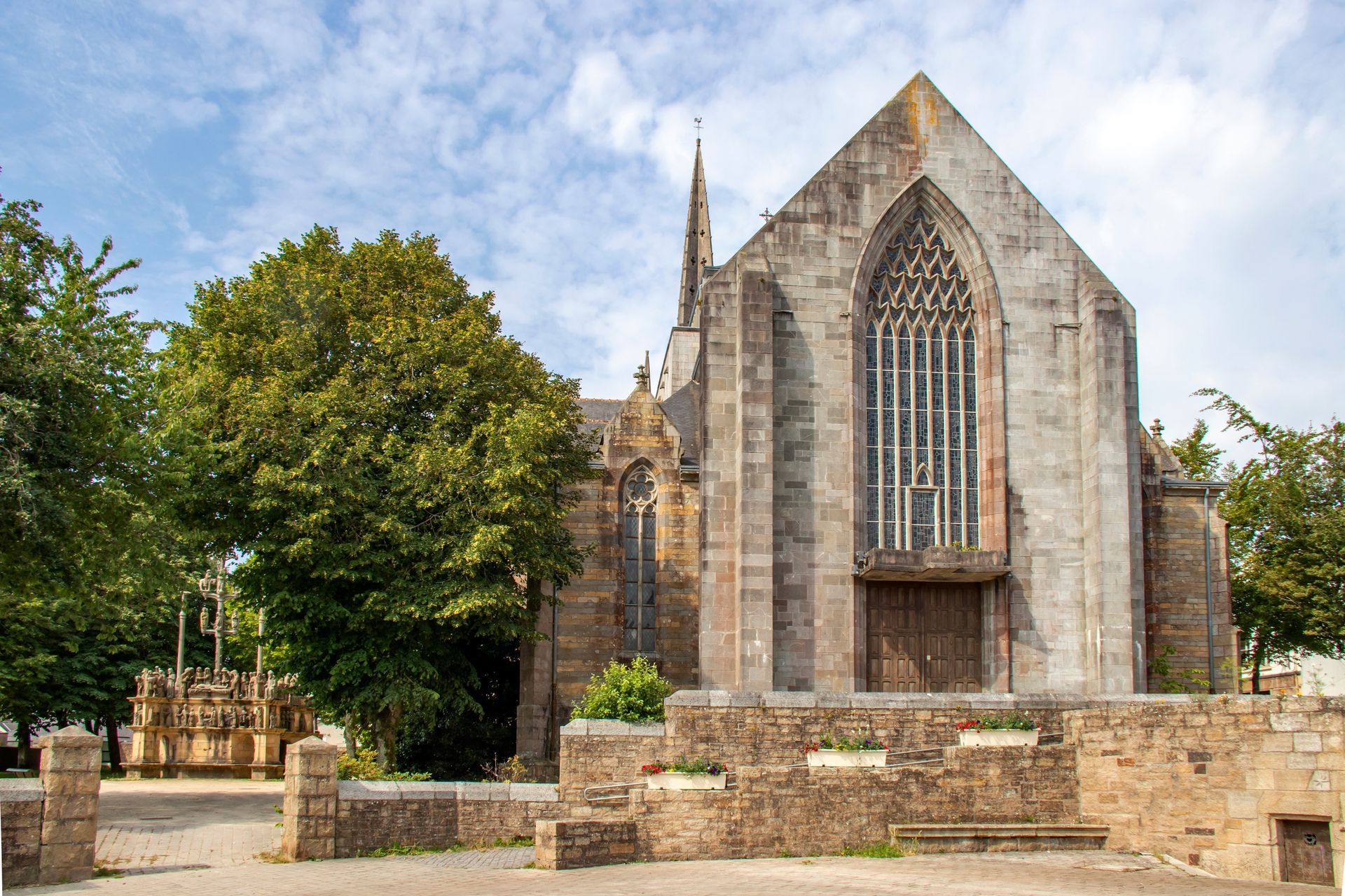 Église en pierre avec une grande fenêtre cintrée, une flèche et des arbres et un mur environnants.