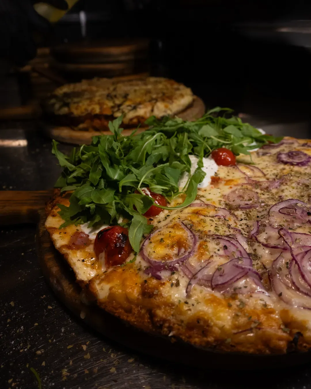 Pizza con cebolla roja y rúcula sobre una tabla de madera. Otra pizza al fondo.
