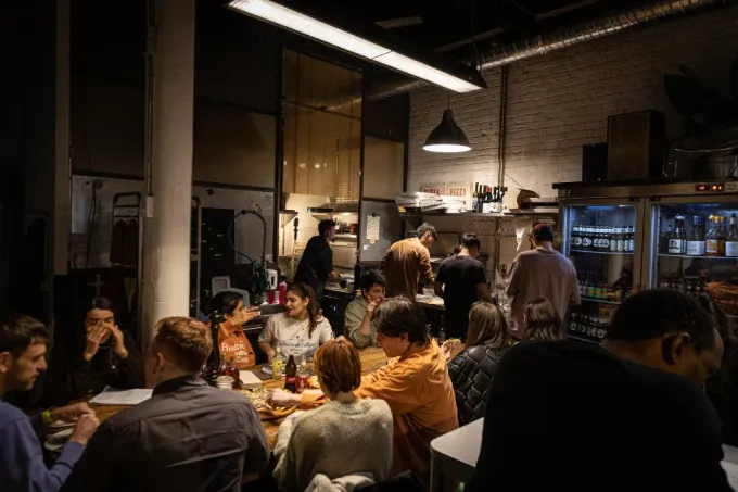 Interior del restaurante; gente sentada en mesas, algunos conversando. Personal de cocina visible al fondo, con poca luz.