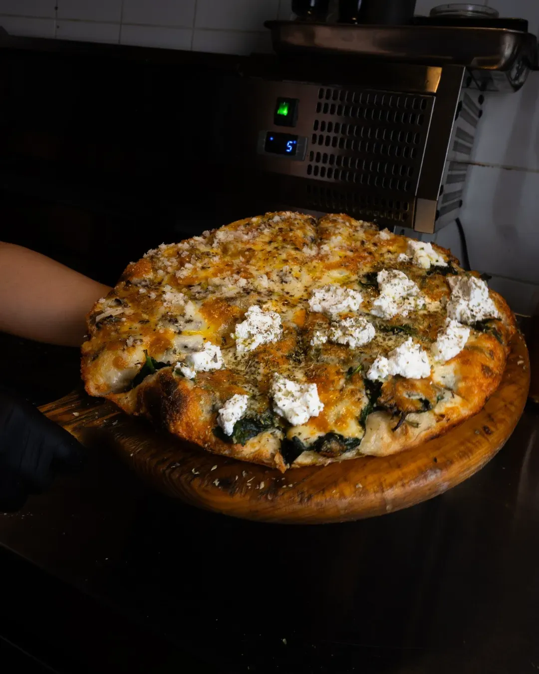 Pizza sobre una pala de madera, cubierta con queso y hierbas, preparada en una cocina.