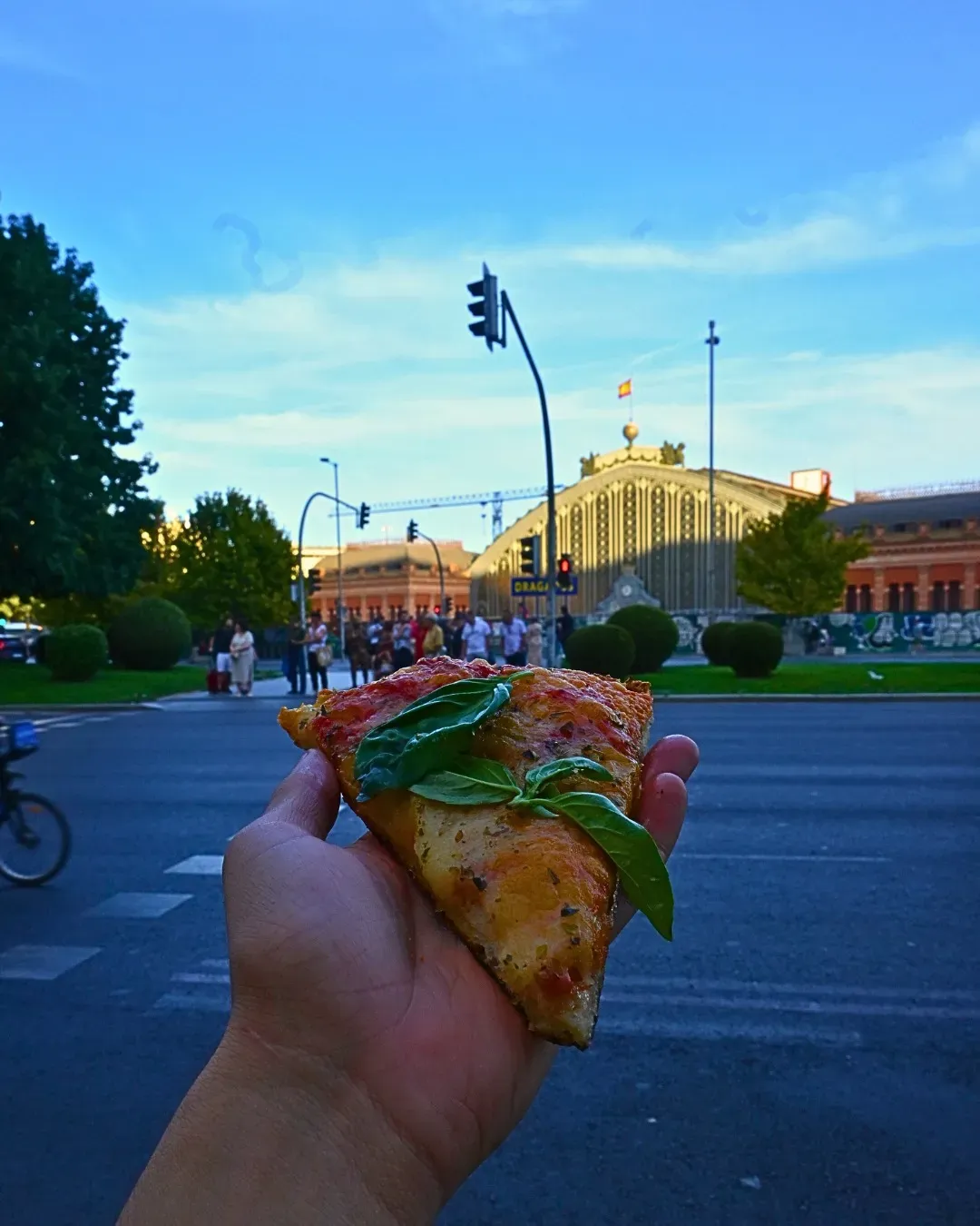 Mano sosteniendo una porción de pizza con albahaca frente a una calle de la ciudad y un edificio bajo un cielo azul.