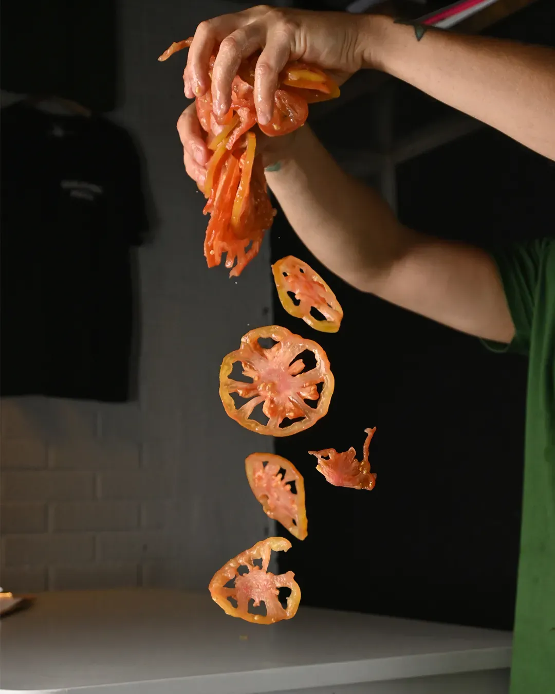 Una persona exprime rodajas de tomate sobre un mostrador blanco. Las rodajas caen. Fondo oscuro.