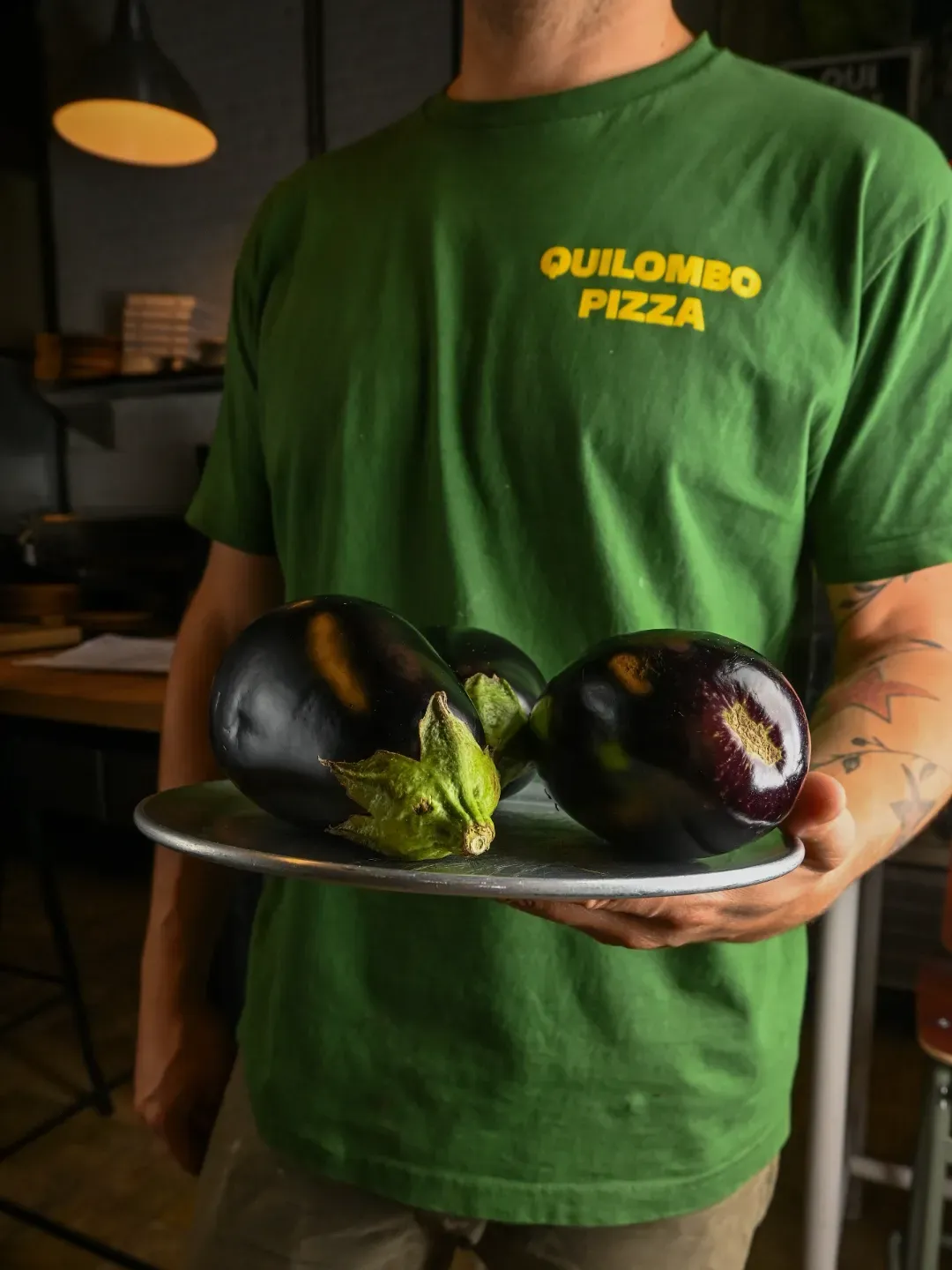 Hombre con camisa verde sosteniendo un plato con tres berenjenas. El logo de Quilombo Pizza está en la camisa.