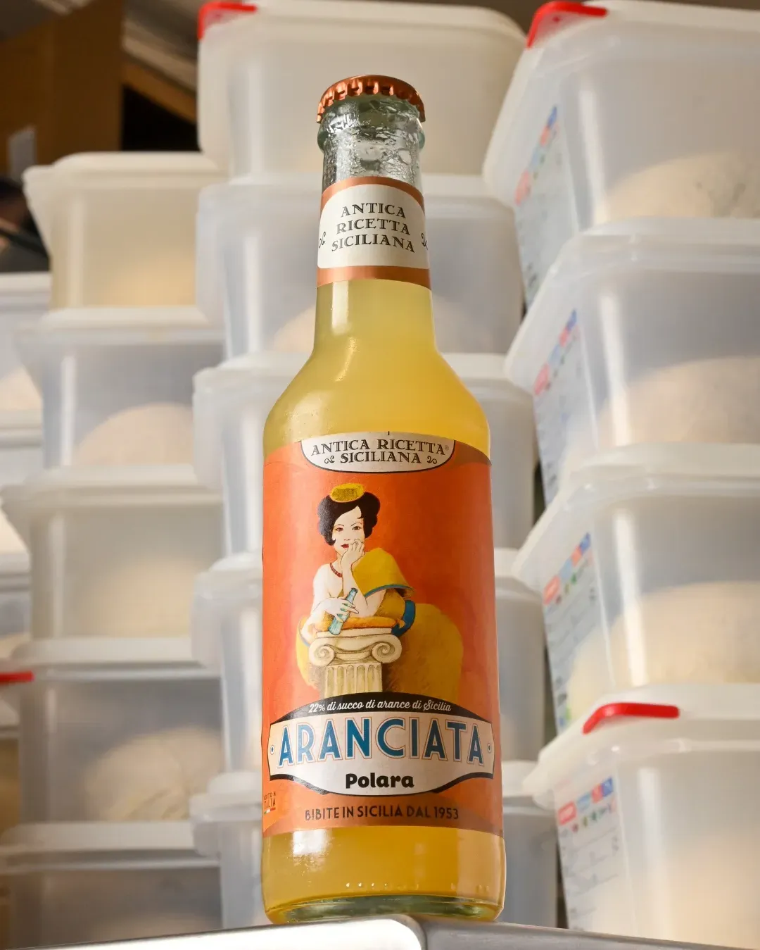 Botella de Aranciata Polara de color naranja con una etiqueta de una mujer y naranjas, delante de contenedores transparentes apilados.