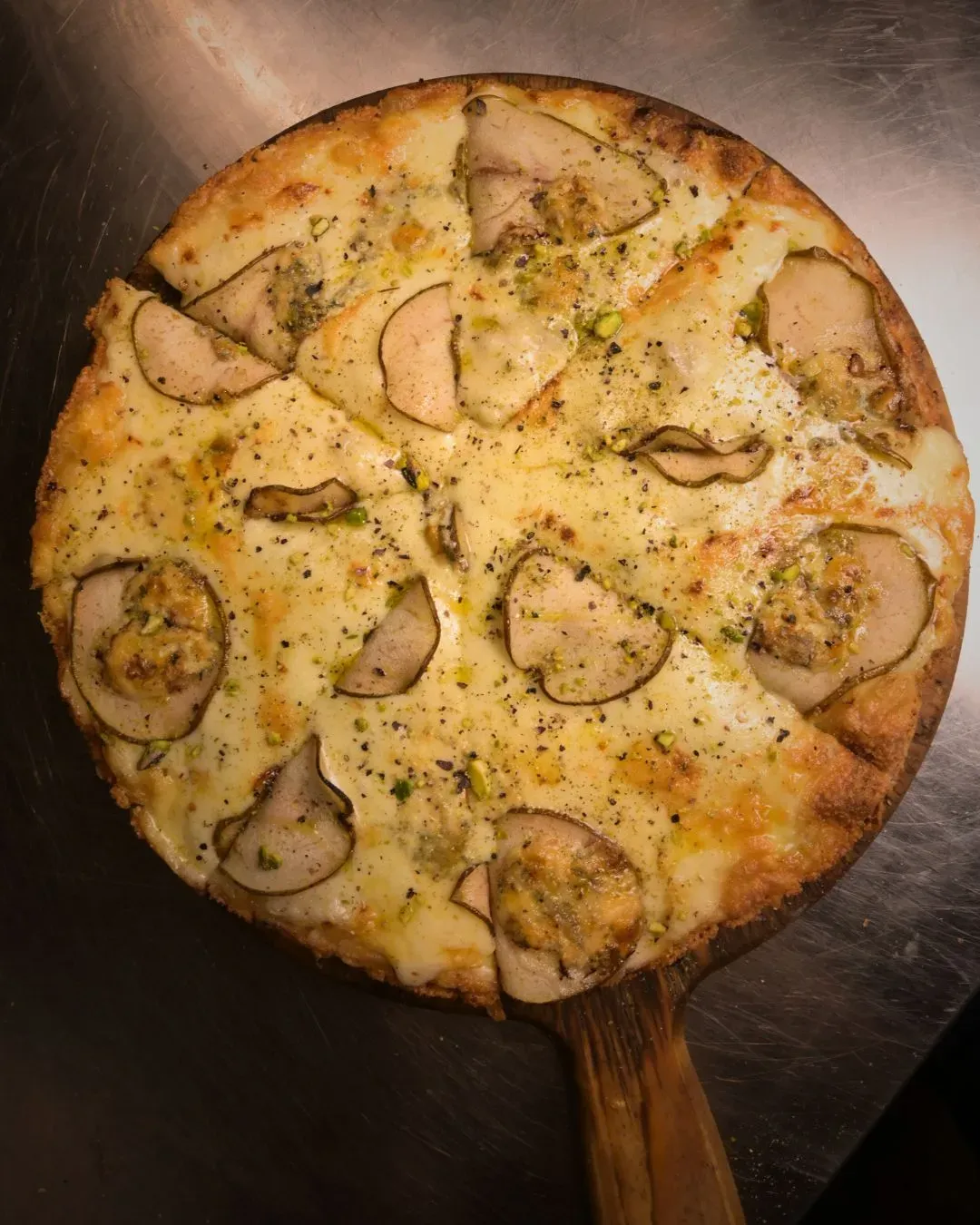 Pizza cubierta con rodajas de pera y motas verdes sobre una paleta de madera, cortada en rebanadas.