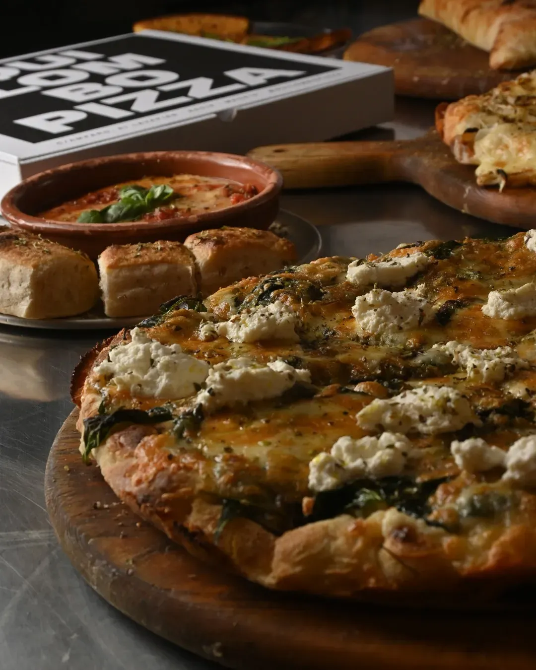 Pizza con ricotta y espinacas, guarniciones y una caja de pizza sobre una mesa.
