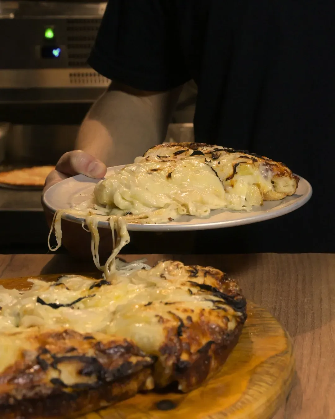 Persona sosteniendo un plato con una porción de pizza con queso, pizza sobre una tabla de madera debajo, horno al fondo.