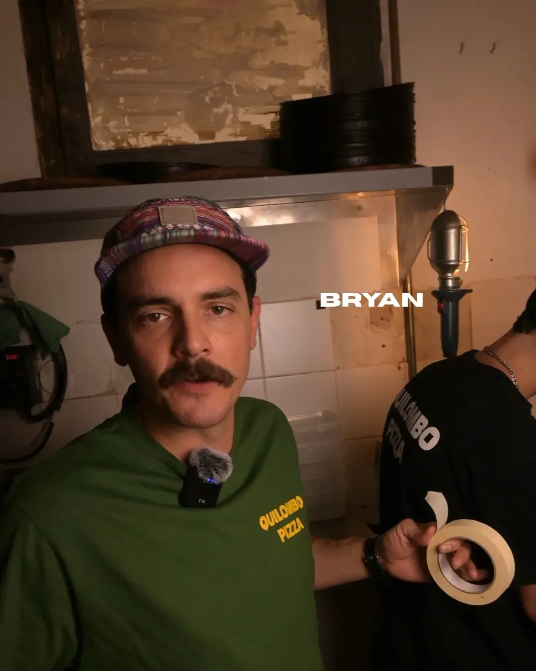 Hombre con camisa verde y gorra con bigote, de pie en una cocina; superposición de