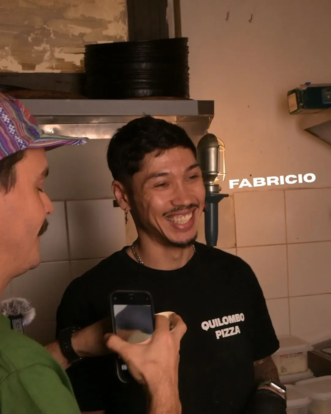 Hombre con camisa negra sonriendo, filmado por otra persona. En la cocina. Etiqueta