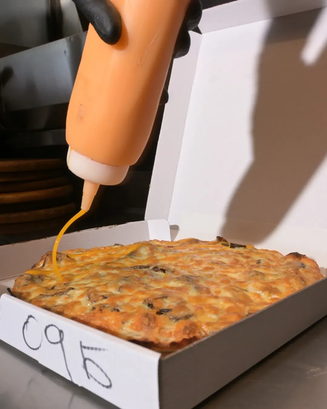 Se vierte salsa sobre una pizza cuadrada en una caja. La pizza tiene la superficie dorada y la etiqueta