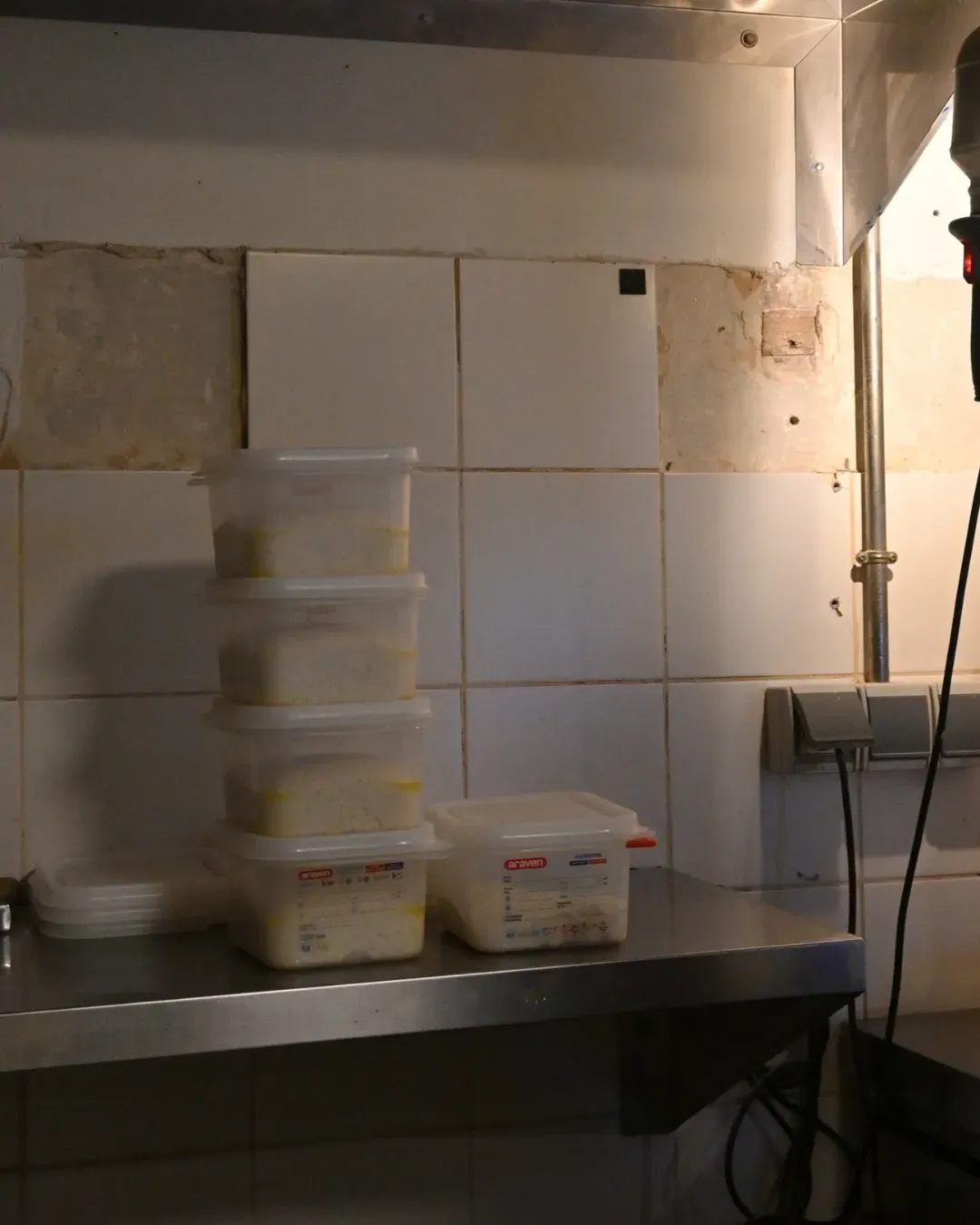 Pila de recipientes de alimentos transparentes sobre un estante de acero inoxidable, contra una pared de azulejos en una cocina.