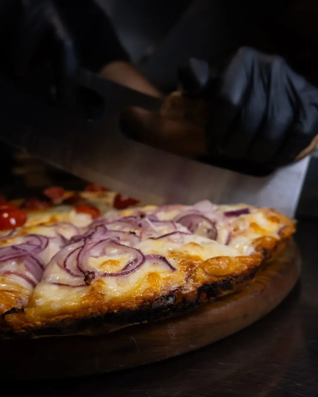 Pizza cortada en rodajas sobre una tabla de madera, cubierta con cebollas rojas y tomates; manos enguantadas en negro.