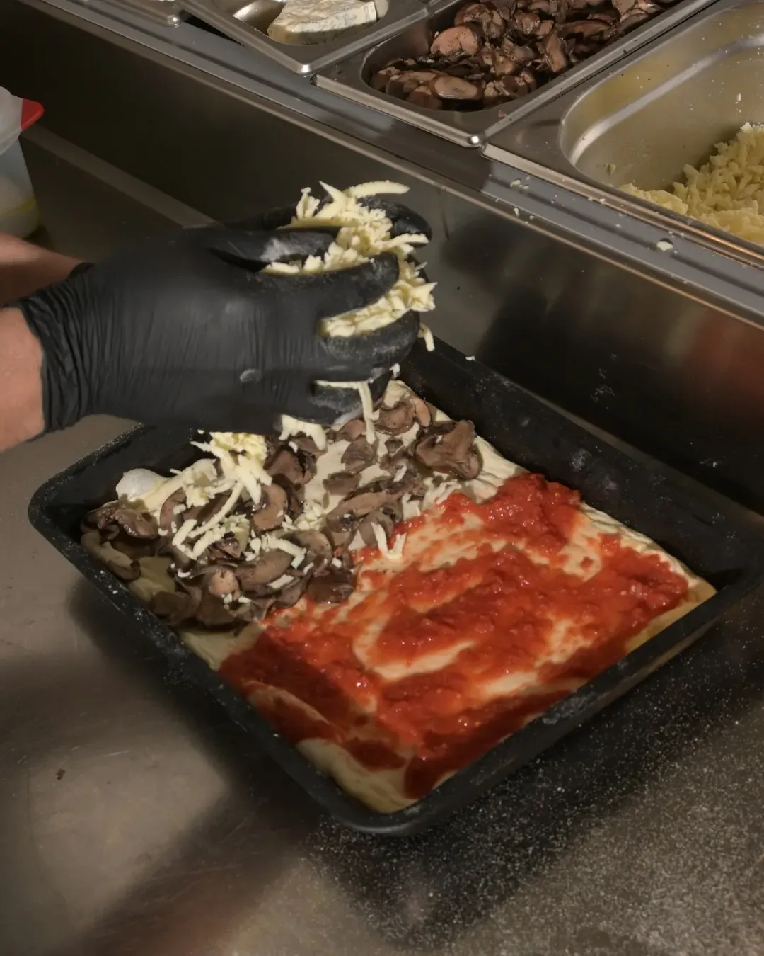 Mano enguantada agregando queso rallado a una base de pizza con salsa y champiñones en un área de preparación de alimentos.