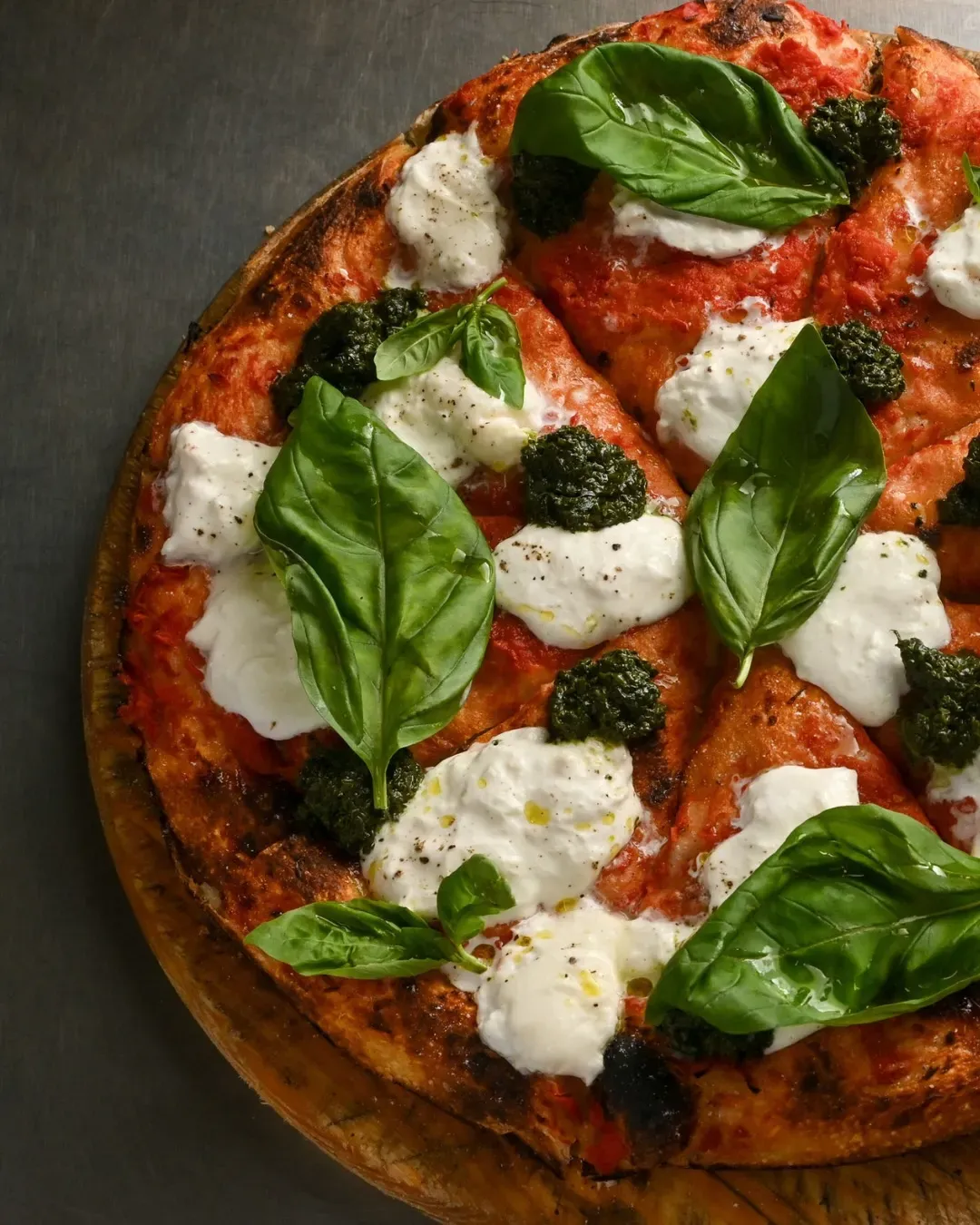 Pizza sobre tabla de madera cubierta con queso blanco, pesto y hojas de albahaca.