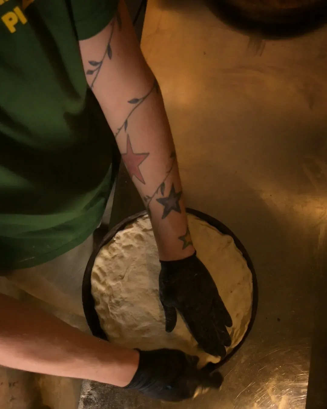 Persona que viste una camisa verde y guantes negros, presionando masa de pizza en una sartén sobre una superficie metálica.