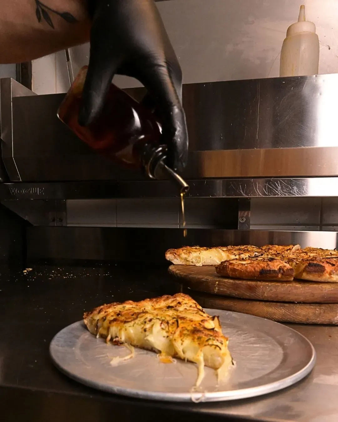 Mano enguantada de negro que vierte miel sobre una rebanada de pizza con queso en una placa de metal, cerca de una tabla de pizza de madera.