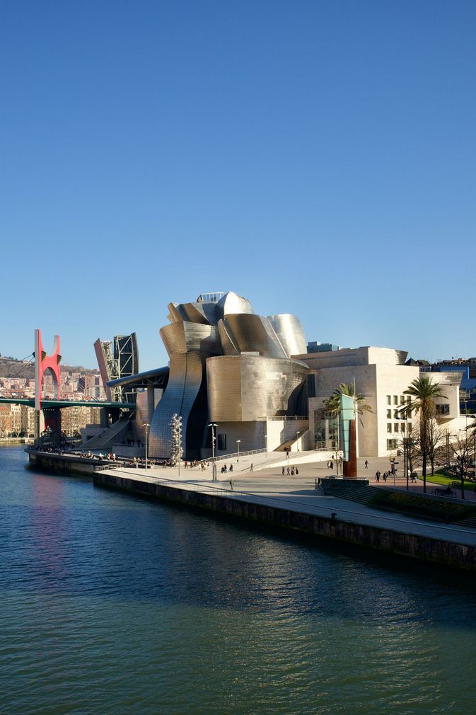 Museo moderno junto a un río bajo un cielo azul despejado, con un puente al fondo.