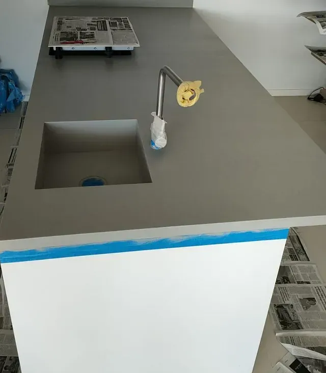 Encimera de cocina con fregadero cuadrado empotrado y grifo moderno. Encimera gris sobre un mueble blanco, con cinta de pintor azul en el borde.