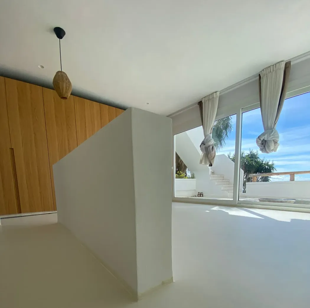 Interior luminoso y moderno con paredes blancas, un armario de madera clara y amplios ventanales con vistas al mar y una escalera. Una singular lámpara colgante cuelga del techo.