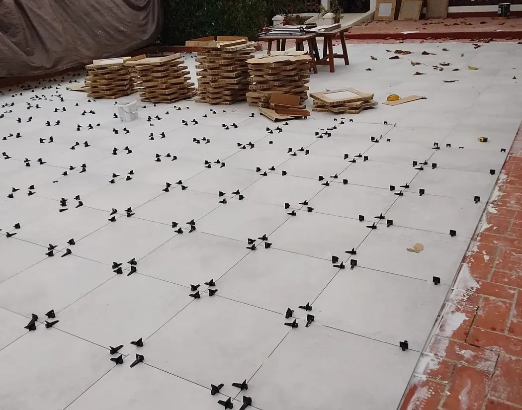 Un suelo de patio con grapas niveladoras de baldosas oscuras que marcan patrones de cuadrícula. Se ven pilas de baldosas y herramientas al fondo.