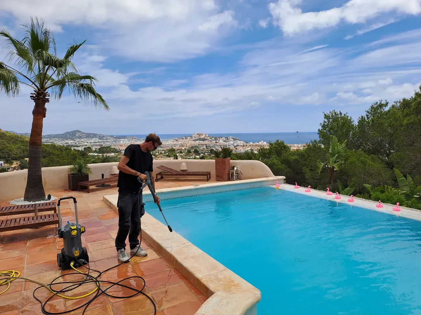 Un hombre limpiando la terraza de una piscina con una hidrolavadora en un día soleado con vistas a una ciudad costera y al mar.