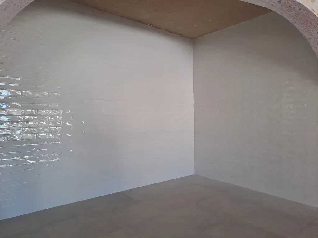 Habitación vacía, de color claro, con paredes blancas y una sección de azulejos a la izquierda. Se aprecia un techo curvo en la parte superior.