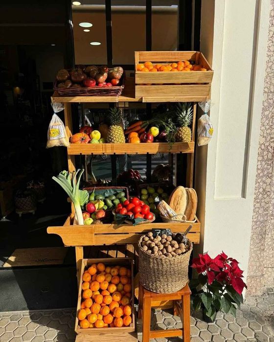 Puesto de frutas de madera en el exterior de una tienda. Exhibe naranjas, piñas, manzanas, puerros y otros productos.