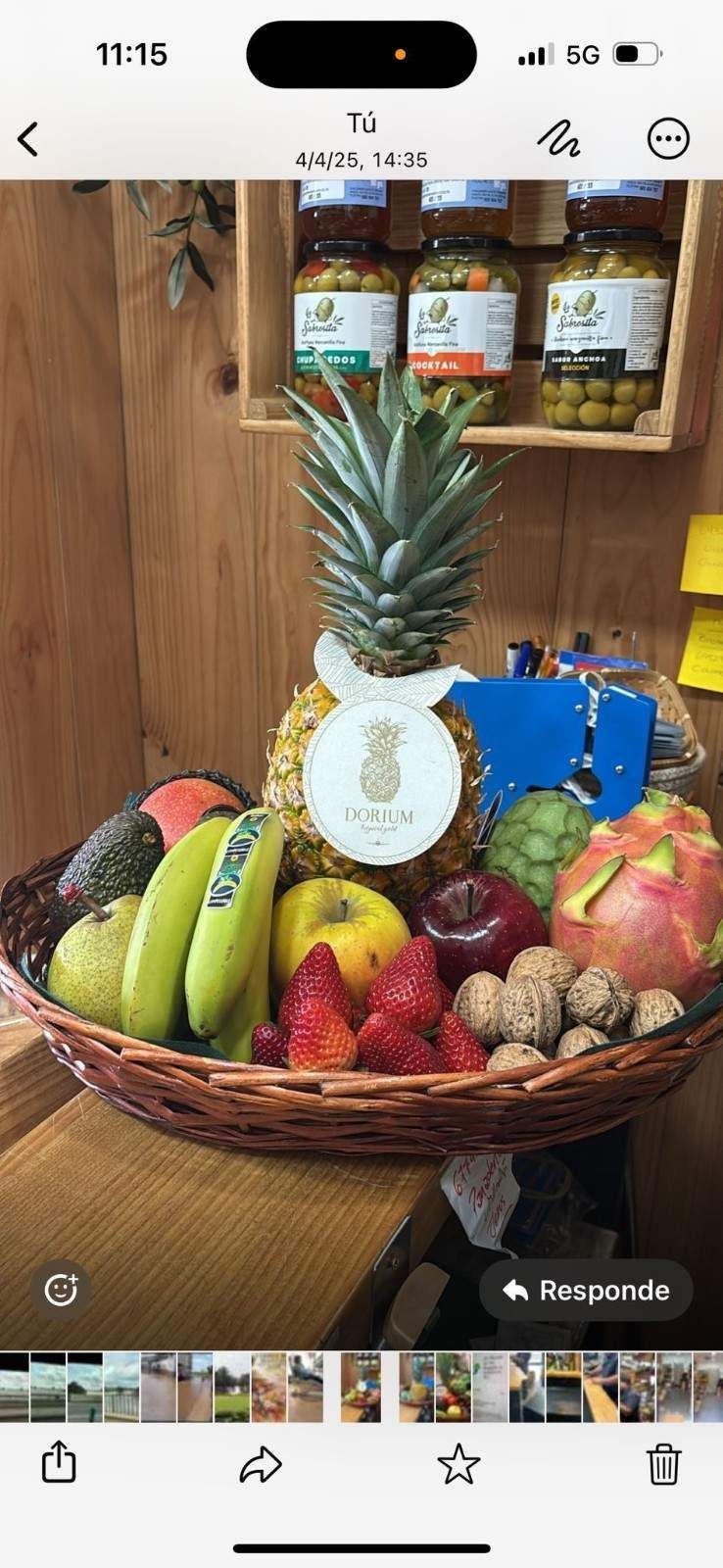 Cesta de frutas con piña, plátanos, fresas, manzanas y nueces; frascos al fondo.