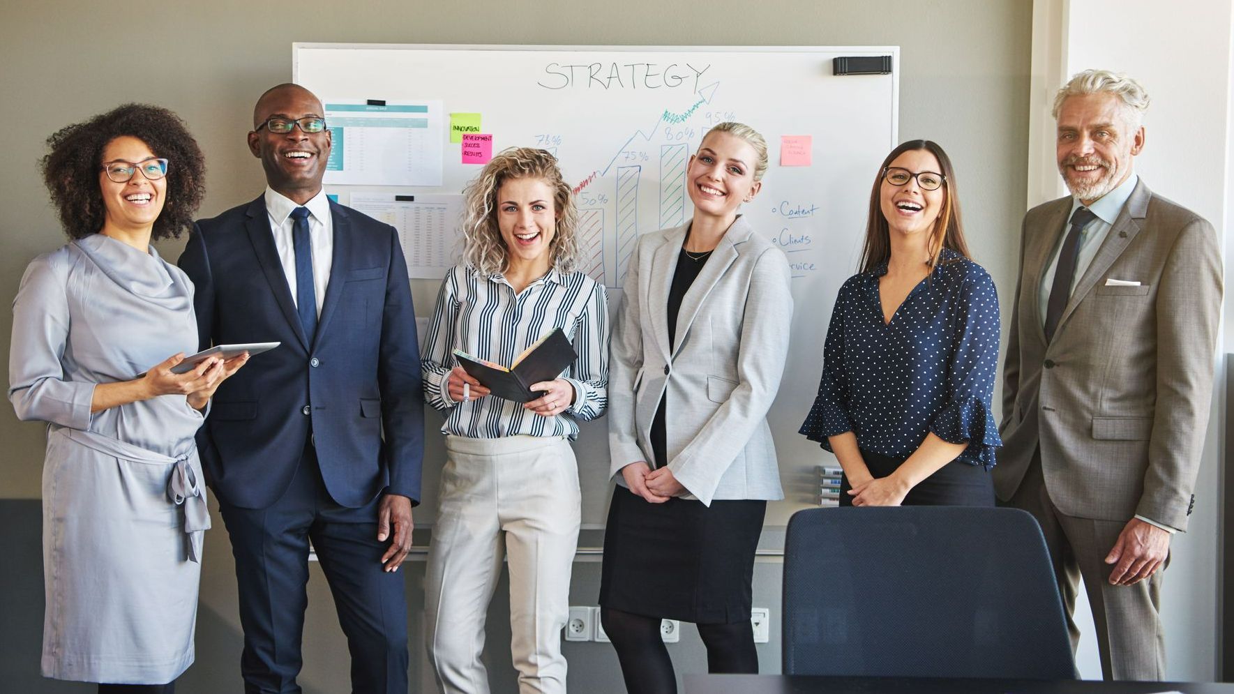 6 collègues qui sourient et posent devant un tableau blanc