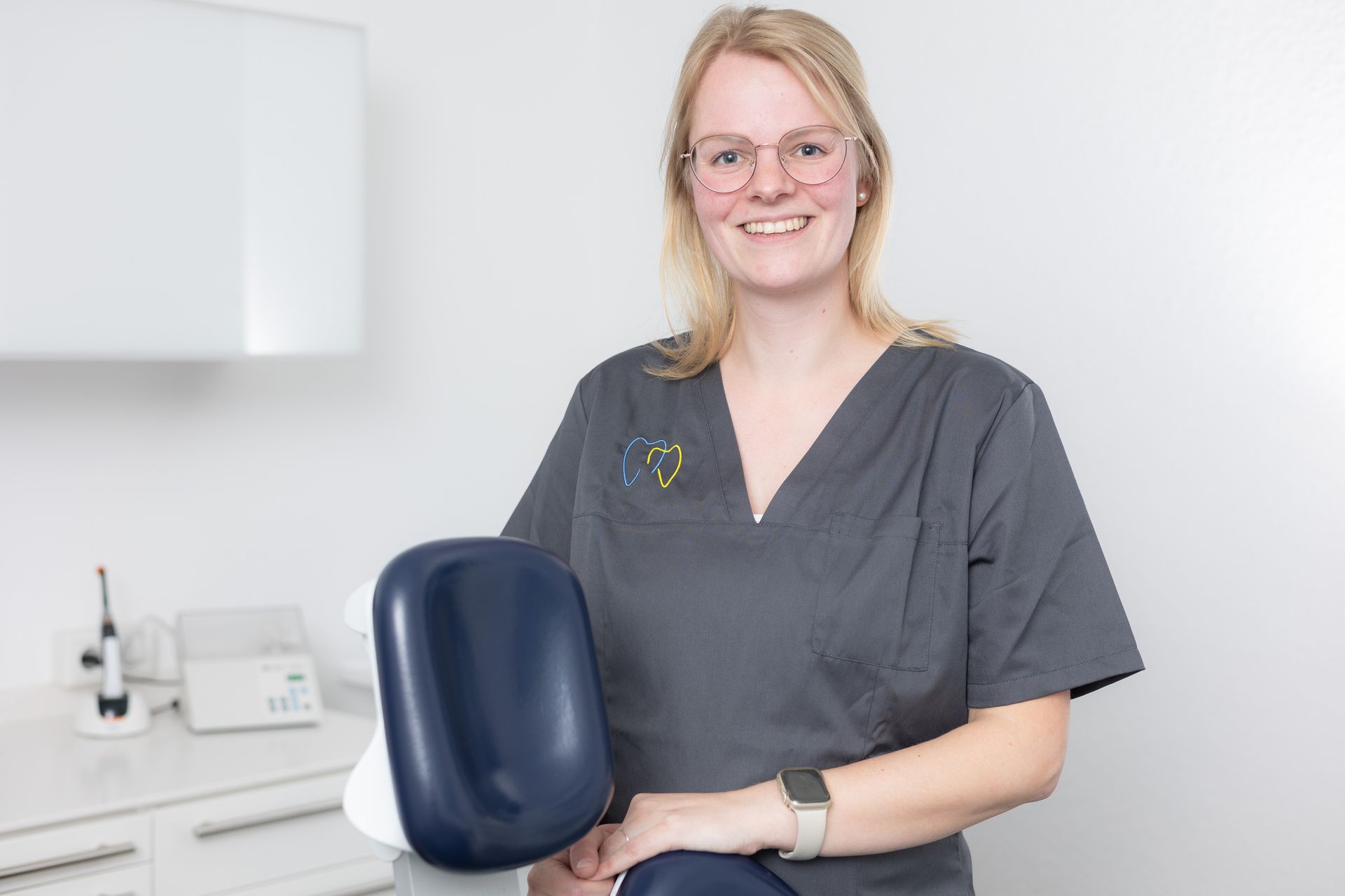Blonde Frau im OP-Kittel, mit Brille, lächelt und hält einen blauen Stuhl in einer Zahnarztpraxis.