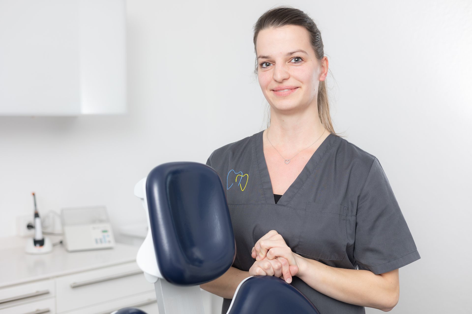 Frau in grauer OP-Kleidung in einer Zahnarztpraxis lächelt und hält einen Zahnarztstuhl.