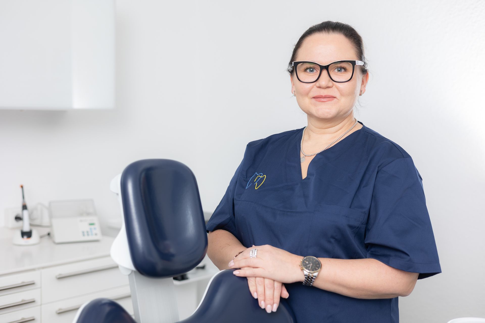 Zahnärztin mit Brille und OP-Kittel, lächelnd, posiert neben einem Zahnarztstuhl in einer Klinik.