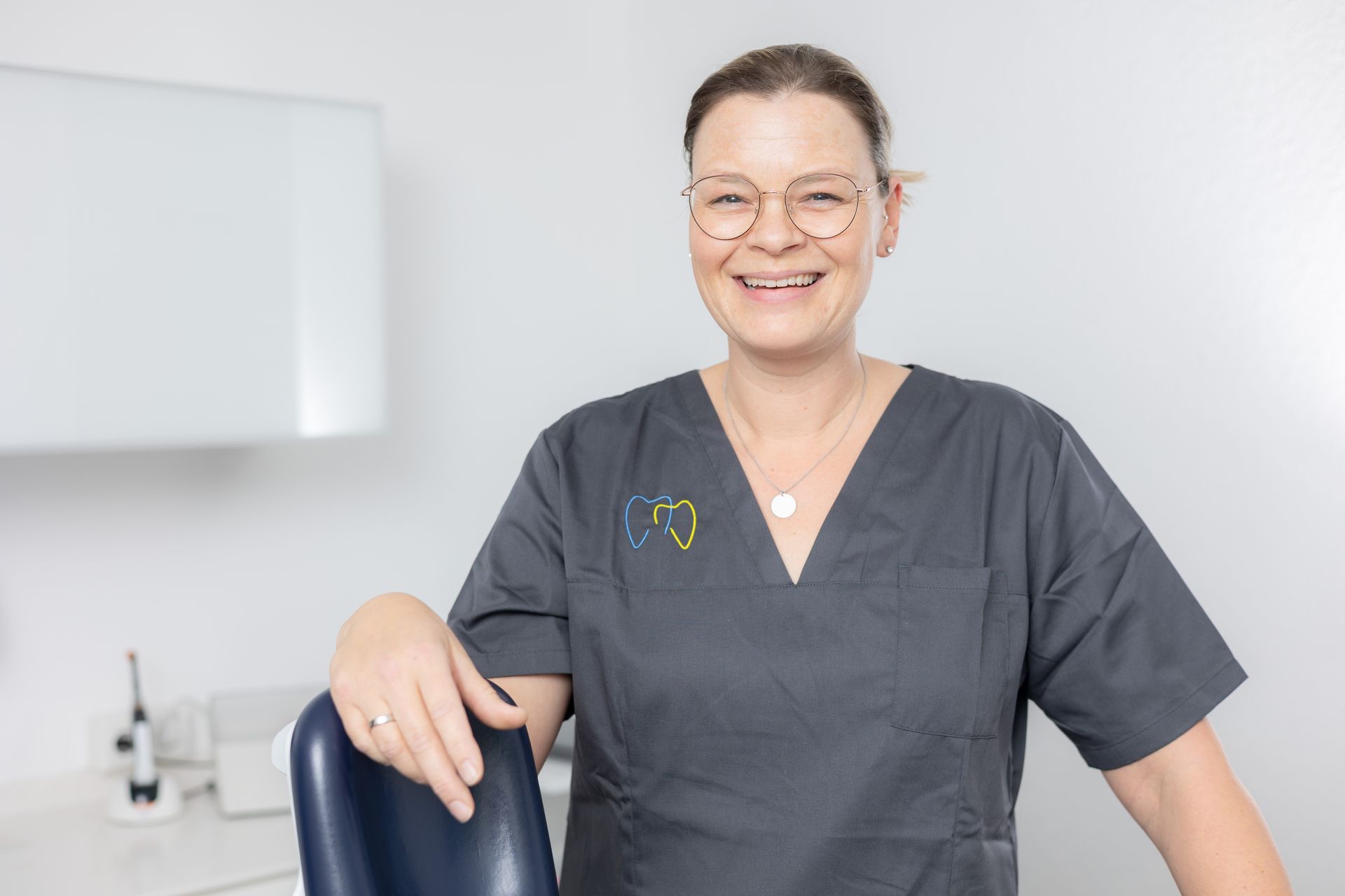 Zahnarzt in grauer OP-Kleidung lächelt in der Praxis und lehnt sich auf einen Stuhl.
