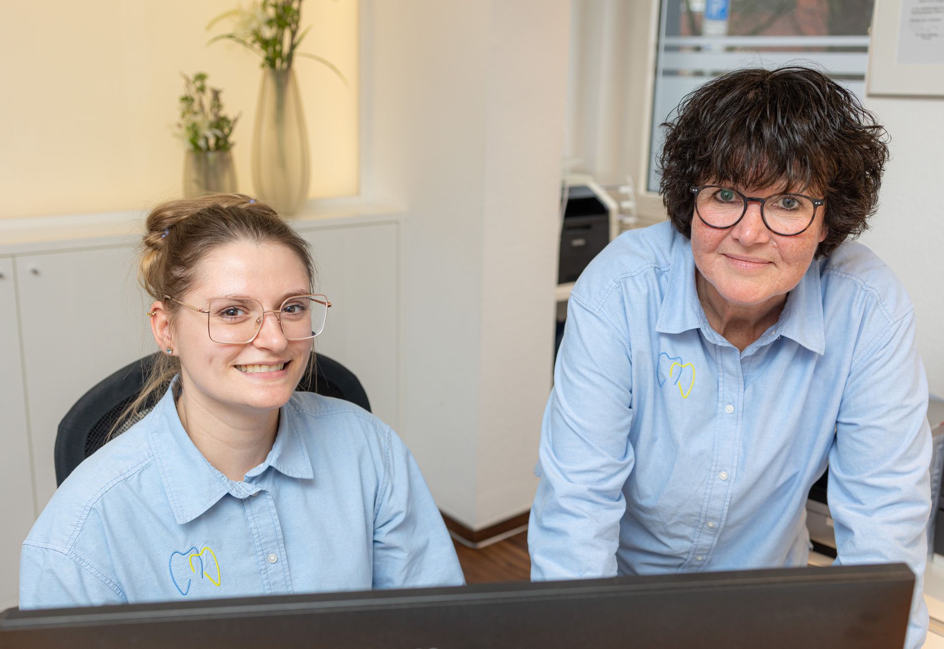 Zwei lächelnde Frauen in hellblauen Hemden und Brillen sitzen in einem Büro vor einem Computer.