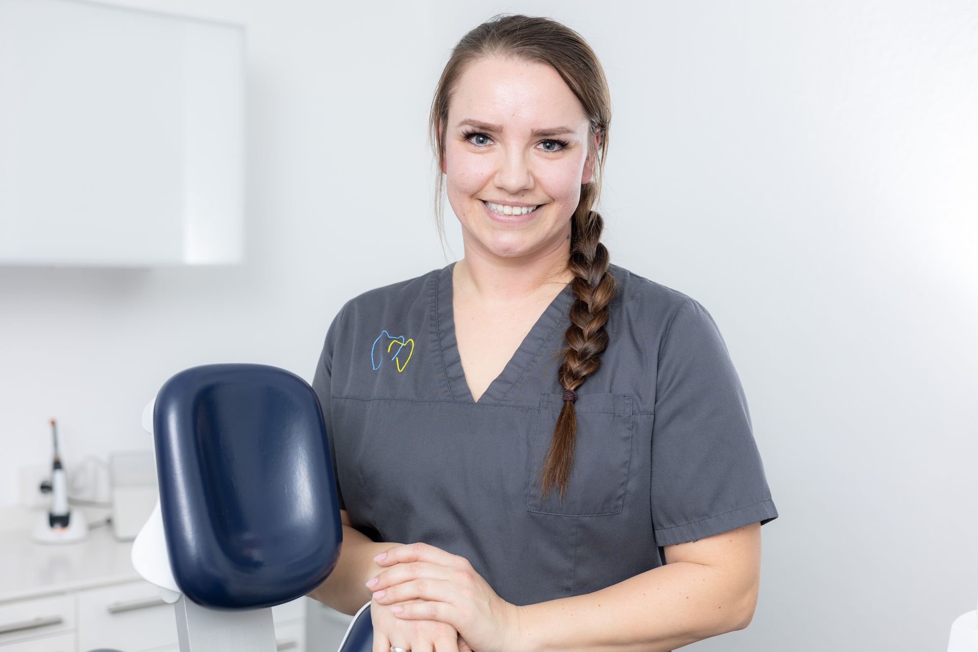 Frau in grauer OP-Kleidung lächelt und lehnt sich in einem hellen Büro auf einen Zahnarztstuhl.