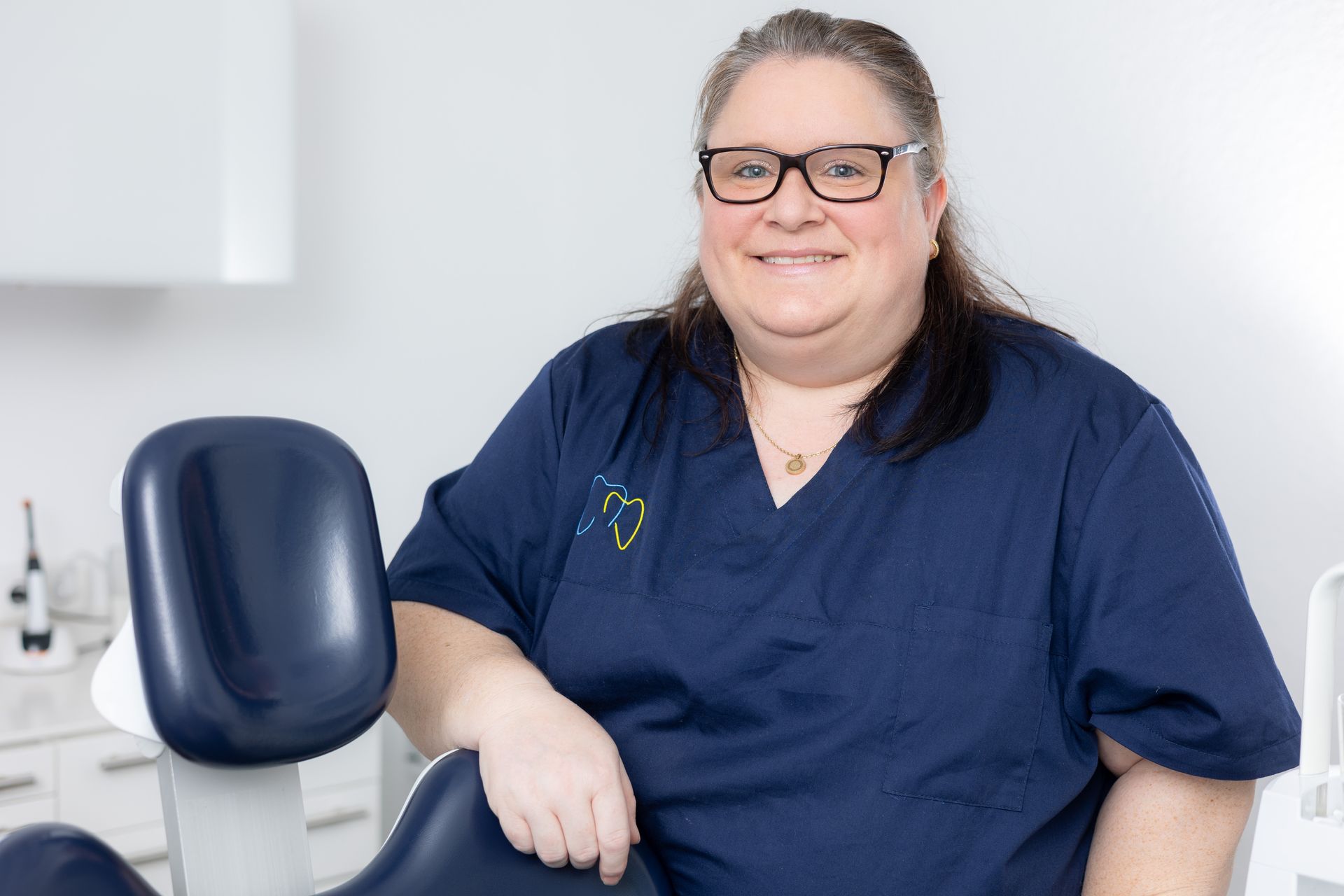 Frau im blauen OP-Kittel lächelt und lehnt sich in einer Klinik auf einen Zahnarztstuhl.