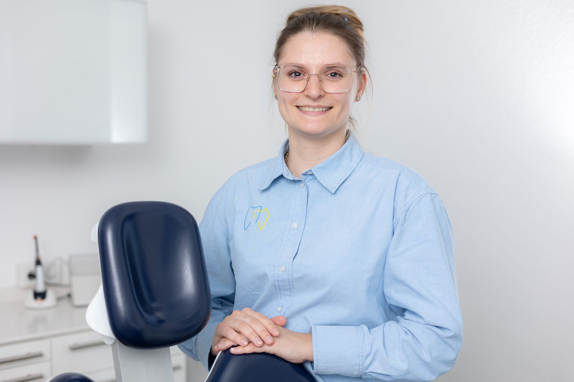 Lächelnde Frau in hellblauem Hemd und Brille steht neben einem Zahnarztstuhl in einem hellen Büro.