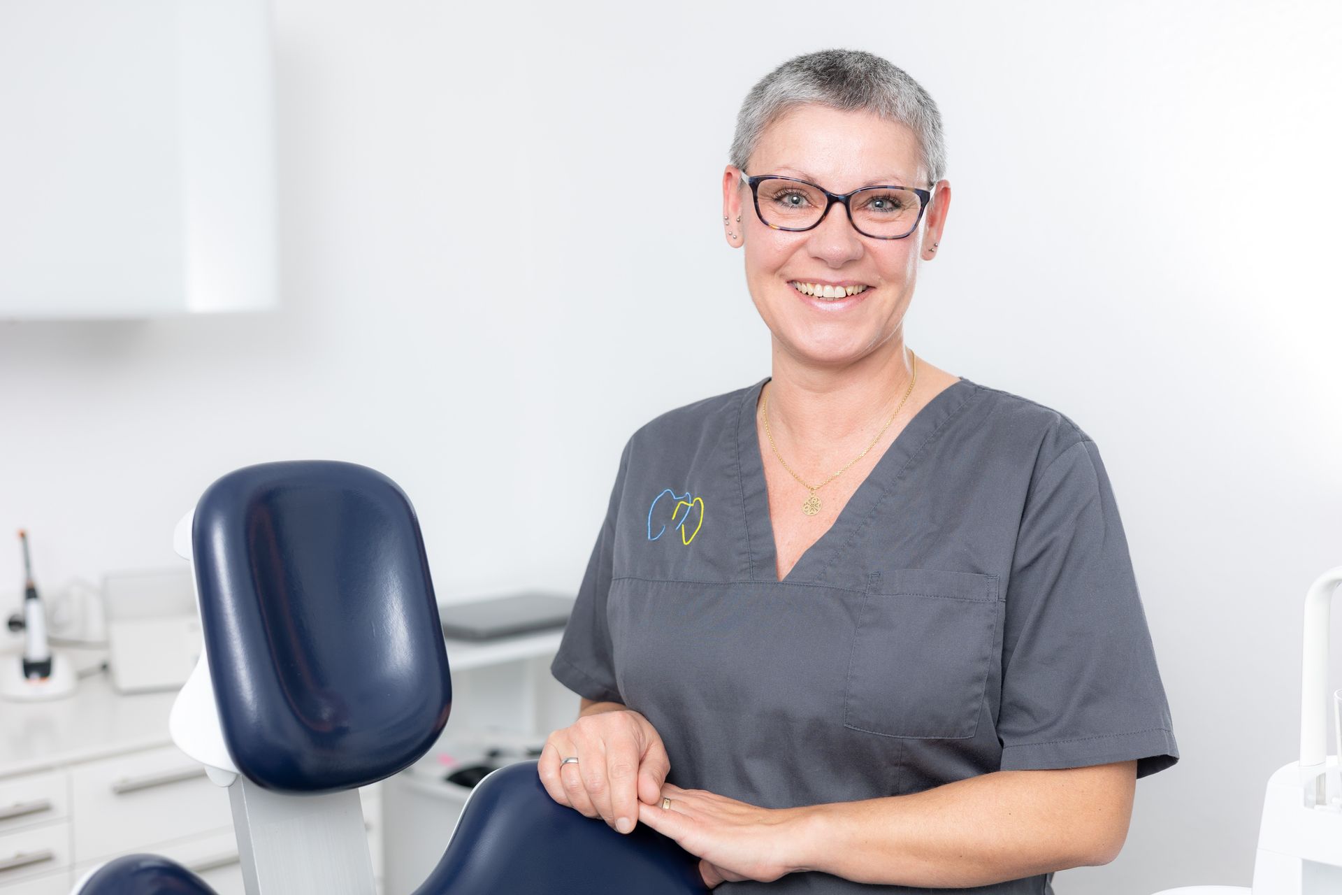 Zahnärztin in grauem Kittel und Brille lächelt in ihrer Praxis.