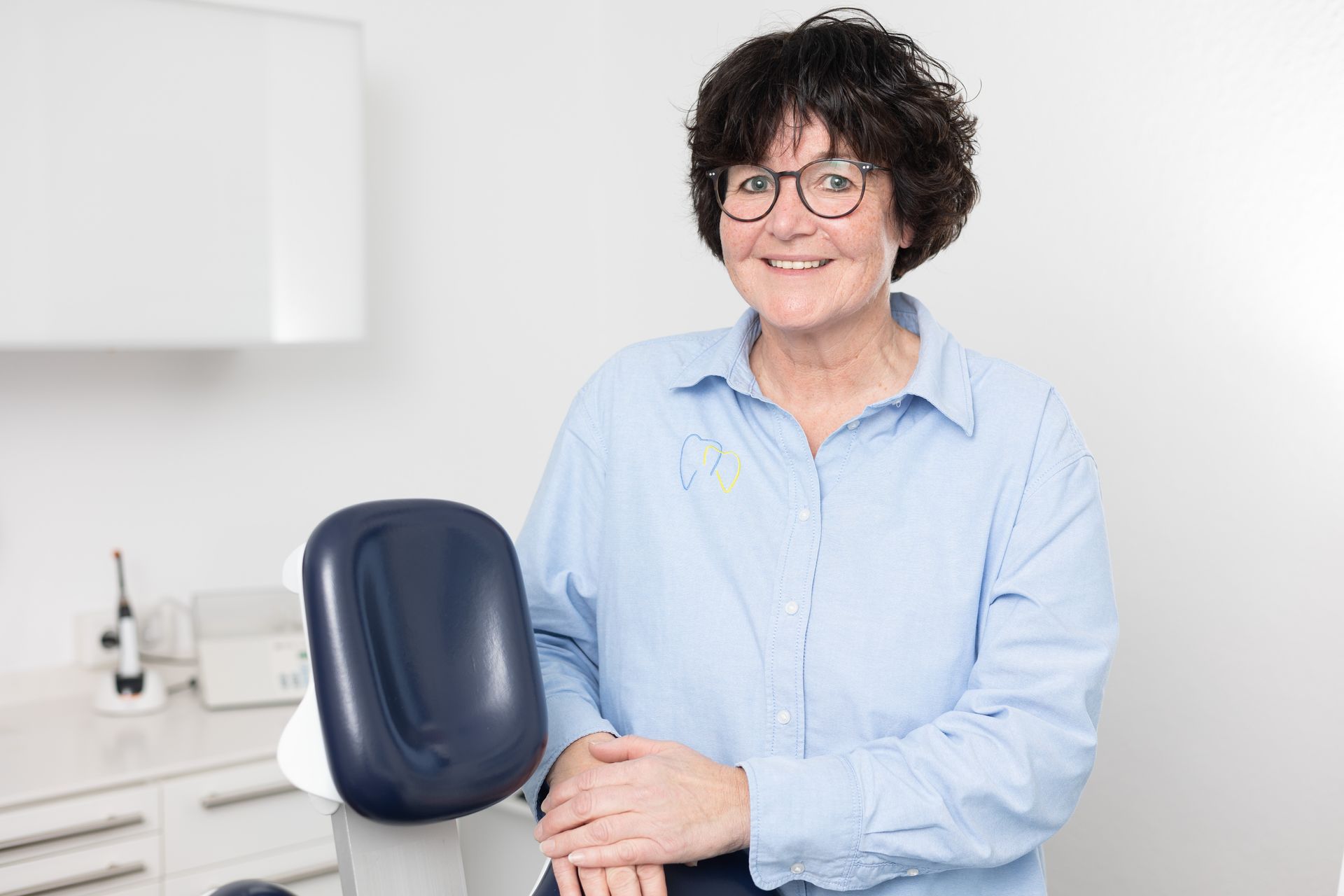 Frau im blauen Hemd lächelt, lehnt auf Zahnarztstuhl, Brille, Klinikumgebung.