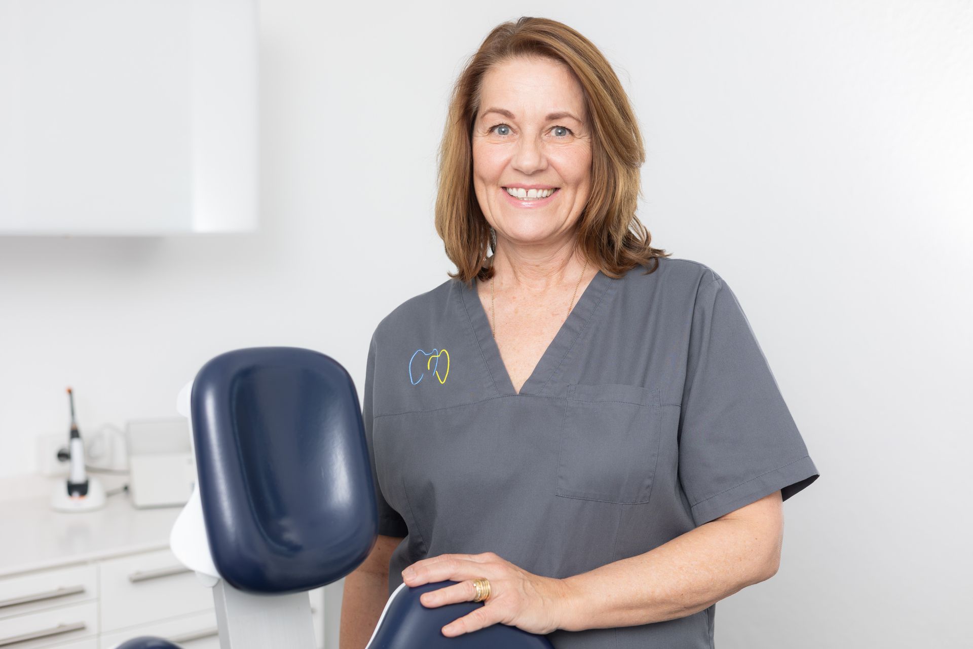 Lächelnde Frau in grauer OP-Kleidung steht in der Zahnarztpraxis neben einem blauen Stuhl.