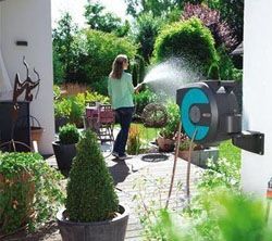 Femme arrosant des plantes dans son jardin avec un tuyau d'arrosage, le dévidoir étant fixé au mur.