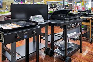 Deux barbecues noirs d'extérieur sont exposés dans un magasin, l'un avec une plaque de cuisson et l'autre avec un couvercle fermé.