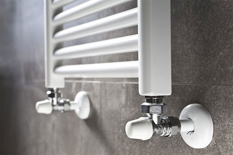 Sèche-serviettes blanc sur un mur carrelé gris, avec robinetterie chromée et cache-robinets blancs.