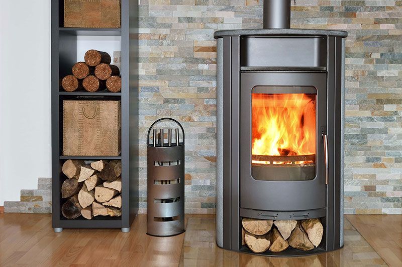 Un poêle à bois avec un feu allumé, du bois de chauffage et une pile de bois contre un mur imitant la pierre.