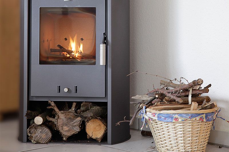 Un poêle à bois gris avec un feu qui brûle, un panier à bois à proximité.