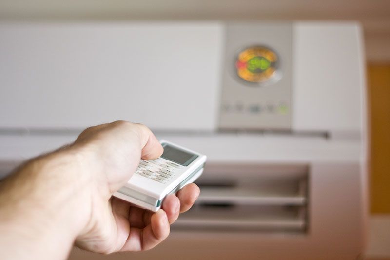 Main tenant une télécommande, pointée vers un climatiseur blanc accroché au mur.