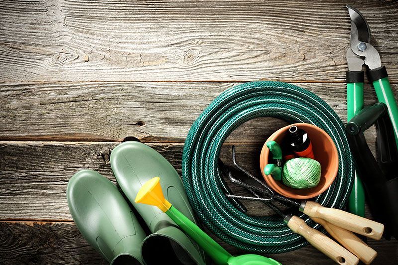 Outils de jardinage sur une surface en bois : tuyau d’arrosage vert, gants, sécateur, truelles, arrosoir et ficelle.