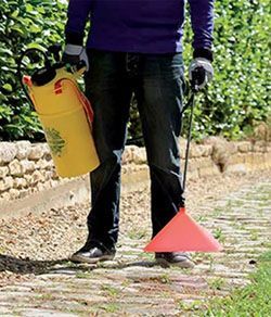 Une personne en jeans tient un pulvérisateur jaune et un déflecteur rose en forme de cône dans un jardin.